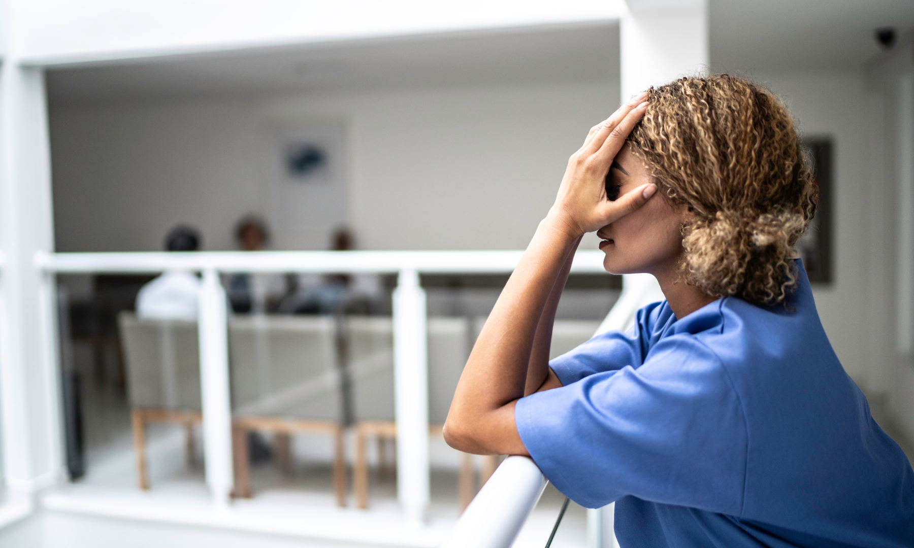 Healthcare professional in blue scrubs leaning on a railing with a concerned expression, hand resting on her forehead.